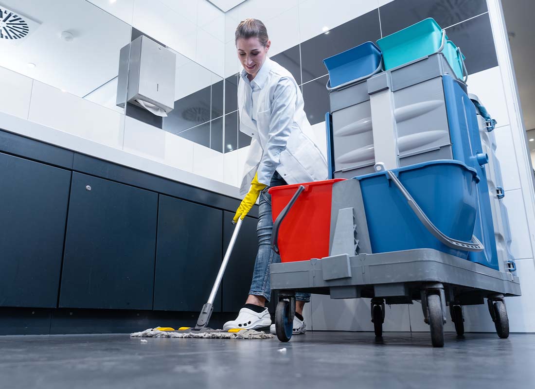 Janitorial Services Insurance - Female Janitor Mopping the Floor of a Restroom With Cleaning Cart Focus and View of Sinks in the Background