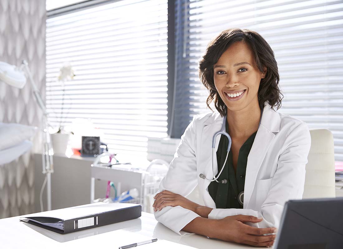 Medical Office Insurance - Portrait of Smiling Female Doctor Wearing White Coat with Stethoscope Sitting behind Her Desk in the Office